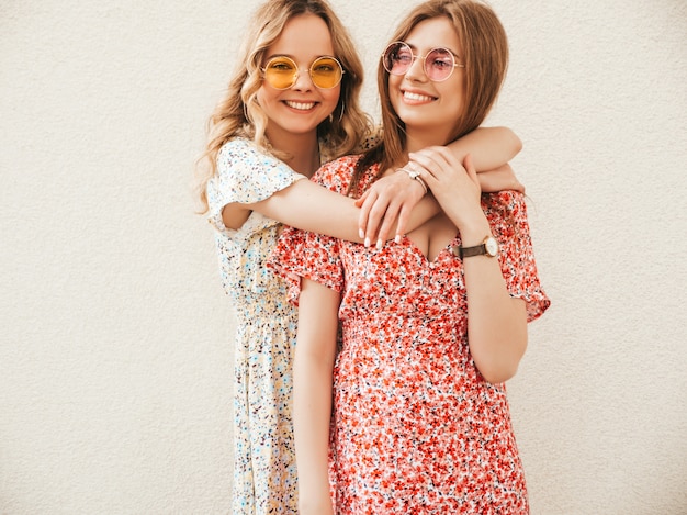 Dos jóvenes hermosas chicas hipster sonrientes en vestido de verano de moda. Mujeres despreocupadas sexy posando junto a la pared en el fondo de la calle en gafas de sol. Modelos positivos divirtiéndose y abrazándose