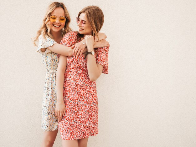 Dos jóvenes hermosas chicas hipster sonrientes en vestido de verano de moda. Mujeres despreocupadas sexy posando junto a la pared en el fondo de la calle en gafas de sol. Modelos positivos divirtiéndose y abrazándose