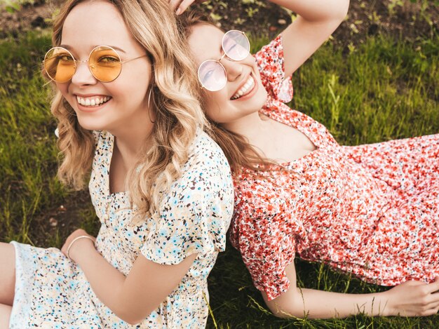 Dos jóvenes hermosas chicas hipster sonrientes en vestido de verano de moda. Mujeres despreocupadas sexys que mienten en la hierba verde en gafas de sol. Modelos positivos que se divierten. Vista superior