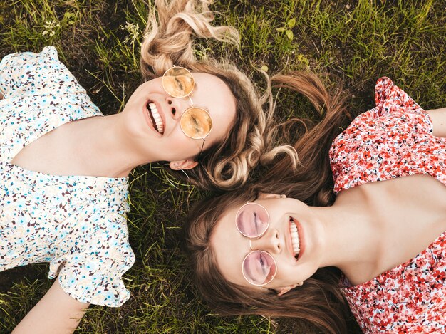 Dos jóvenes hermosas chicas hipster sonrientes en vestido de verano de moda. Mujeres despreocupadas sexys que mienten en la hierba verde en gafas de sol. Modelos positivos que se divierten. Vista superior