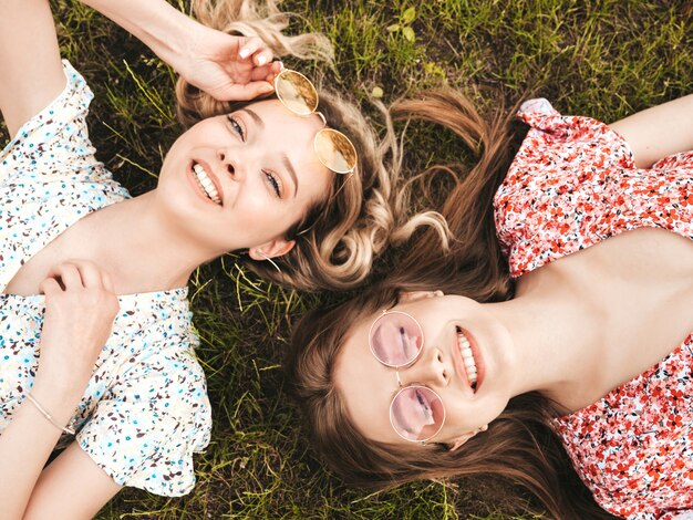 Dos jóvenes hermosas chicas hipster sonrientes en vestido de verano de moda. Mujeres despreocupadas sexys que mienten en la hierba verde en gafas de sol. Modelos positivos que se divierten. Vista superior