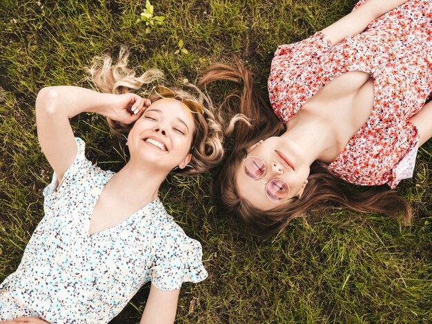 Dos jóvenes hermosas chicas hipster sonrientes en vestido de verano de moda. Mujeres despreocupadas sexys que mienten en la hierba verde en gafas de sol. Modelos positivos que se divierten. Vista superior