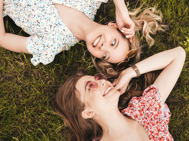 Dos jóvenes hermosas chicas hipster sonrientes en vestido de verano de moda. Mujeres despreocupadas sexys que mienten en la hierba verde en gafas de sol. Modelos positivos que se divierten. Vista superior