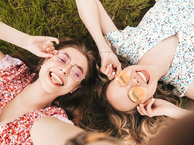 Dos jóvenes hermosas chicas hipster sonrientes en vestido de verano de moda. Mujeres despreocupadas y sexys tumbadas en la hierba verde con gafas de sol. Modelos positivos divirtiéndose. Vista superior. Tomando fotos selfie en smartphone
