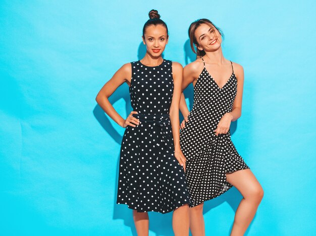 Dos jóvenes hermosas chicas hipster sonrientes en vestidos de moda de lunares de verano. Mujeres despreocupadas sexy posando junto a la pared azul. Divertirse y abrazarse. Modelos muestra buena relación. Mujer con labios rojos