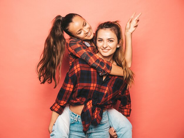 Dos jóvenes hermosas chicas sonrientes morenas hipster en camisa a cuadros de moda similar y ropa de jeans. Mujeres despreocupadas sexy posando cerca de la pared azul en el estudio. Modelos positivos divirtiéndose