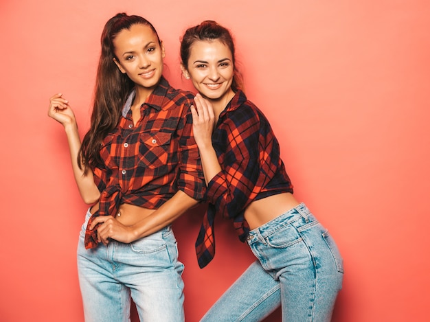 Dos jóvenes hermosas chicas sonrientes morenas hipster en camisa de cuadros similares de moda y ropa de jeans. mujeres despreocupadas sexy posando junto a la pared rosa en el estudio. modelos positivos divirtiéndose