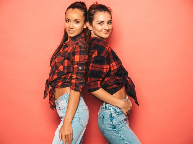 Dos jóvenes hermosas chicas sonrientes morenas hipster en camisa de cuadros similares de moda y ropa de jeans. Mujeres despreocupadas sexy posando junto a la pared rosa en el estudio. Modelos positivos divirtiéndose