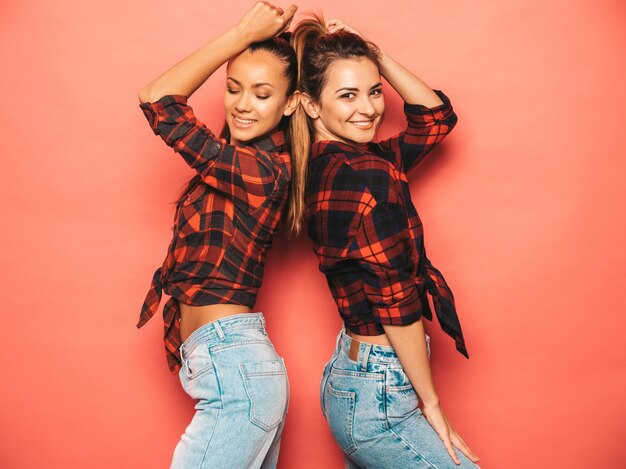 Dos jóvenes hermosas chicas sonrientes morenas hipster en camisa de cuadros similares de moda y ropa de jeans. Mujeres despreocupadas sexy posando junto a la pared rosa en el estudio. Modelos positivos divirtiéndose