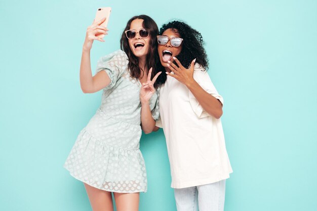 Dos jóvenes hermosas mujeres hipster internacionales sonrientes en ropa de verano de moda Mujeres sexy despreocupadas posando junto a la pared azul en el estudio Modelos positivos divirtiéndose en gafas de sol Tomando fotos selfie