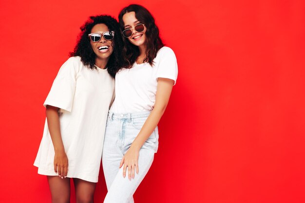 Dos jóvenes hermosas mujeres hipster internacionales sonrientes en ropa de verano de moda Mujeres sexy despreocupadas posando junto a la pared roja en el estudio Modelos positivos divirtiéndose Concepto de amistad En gafas de sol