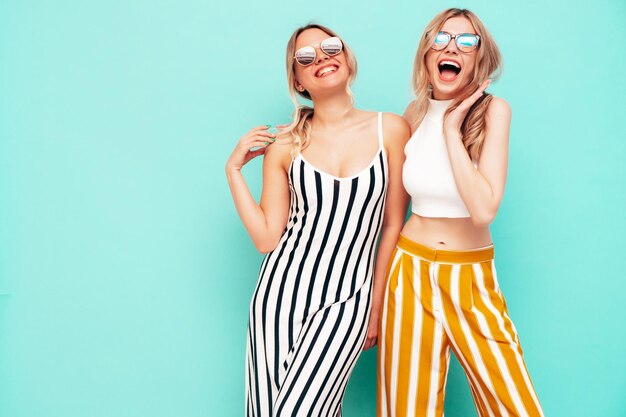 Dos jóvenes hermosas mujeres hipster morena sonriente en ropa de verano de moda Mujeres sexy despreocupadas posando junto a la pared azul en el estudio Modelos positivos divirtiéndose Alegre y feliz