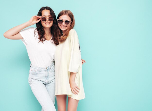 Dos jóvenes hermosas mujeres hipster morena sonriente en ropa de verano de moda Mujeres sexy despreocupadas posando junto a la pared azul Modelos positivos divirtiéndose Alegre y feliz En gafas de sol