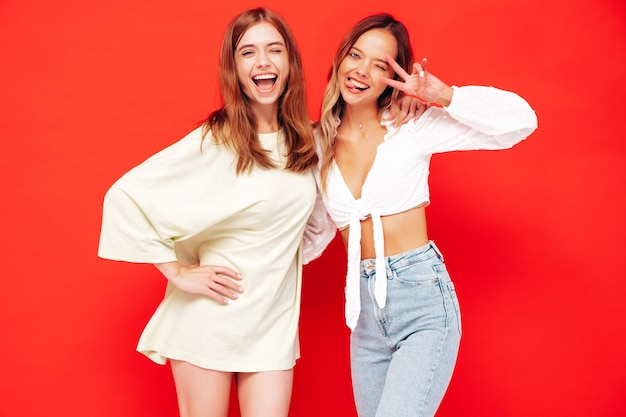 Dos jóvenes hermosas mujeres hipster morena sonriente en ropa de verano de moda Mujeres sexy despreocupadas posando junto a la pared roja Modelos positivos divirtiéndose Alegre y feliz Muestra el signo de la paz