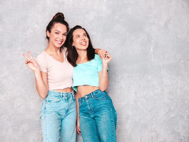 Dos jóvenes hermosas mujeres hipster rubias sonrientes en ropa de verano de moda mujeres despreocupadas posando cerca de la pared gris en el estudio Modelos positivos divirtiéndose Alegre y feliz