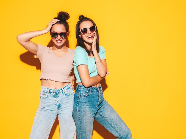 Dos jóvenes hermosas mujeres hipster rubias sonrientes en ropa de verano de moda Mujeres sexy despreocupadas posando junto a la pared amarilla en el estudio Modelos positivos divirtiéndose Alegres y felices En gafas de sol