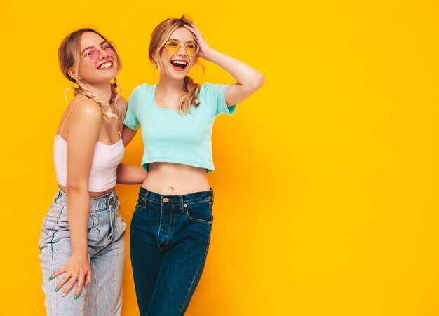 Dos jóvenes hermosas mujeres hipster rubias sonrientes en ropa de verano de moda Mujeres sexy despreocupadas posando junto a la pared amarilla en el estudio Modelos positivos divirtiéndose Alegres y felices En gafas de sol