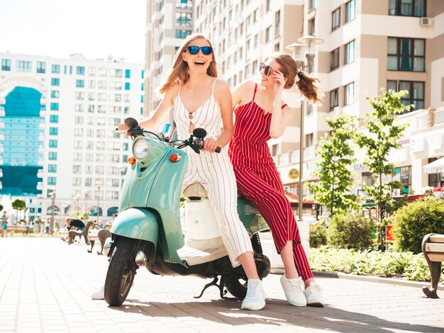 Dos jóvenes hermosas mujeres hipster sonrientes en overoles de moda Mujeres sexy despreocupadas conduciendo moto retro en el fondo de la calle Modelos positivos divirtiéndose montando scooter italiano clásico en anteojos