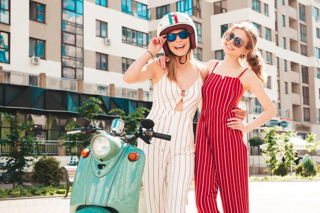 Dos jóvenes hermosas mujeres hipster sonrientes en overoles de moda Mujeres sexy despreocupadas conduciendo moto retro en el fondo de la calle Modelos positivos divirtiéndose montando scooter italiano clásico en anteojos