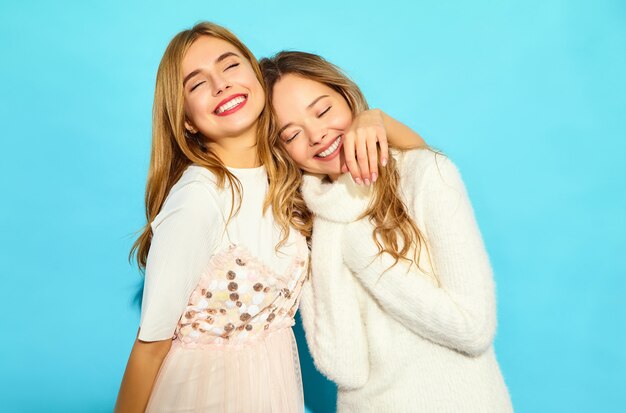 Dos jóvenes hermosas mujeres hipster sonrientes en ropa de moda verano blanco. Mujeres despreocupadas sexy posando junto a la pared azul. Modelos positivos abrazando