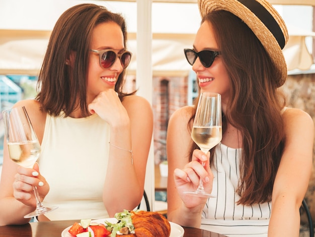 Dos jóvenes hermosas mujeres hipster sonrientes con ropa de verano de moda Mujeres despreocupadas posando en el café de la terraza en la calle Modelos positivos bebiendo vid blanca con sombrero Disfrutando de sus vacaciones