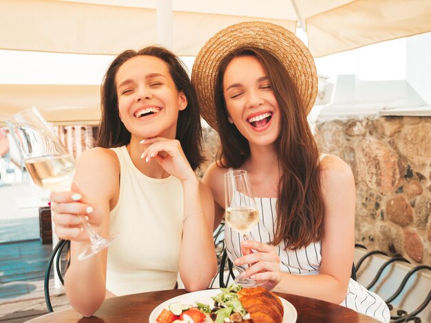 Dos jóvenes hermosas mujeres hipster sonrientes con ropa de verano de moda Mujeres despreocupadas posando en el café de la terraza en la calle Modelos positivos bebiendo vid blanca con sombrero Disfrutando de sus vacaciones