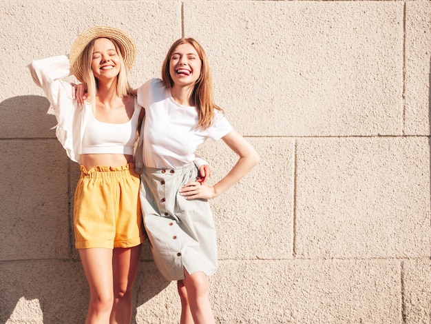 Dos jóvenes hermosas mujeres hipster sonrientes con ropa de verano de moda. Mujeres sexy despreocupadas posando en la calle cerca de una pared blanca con sombrero Modelos puros positivos divirtiéndose al atardecer