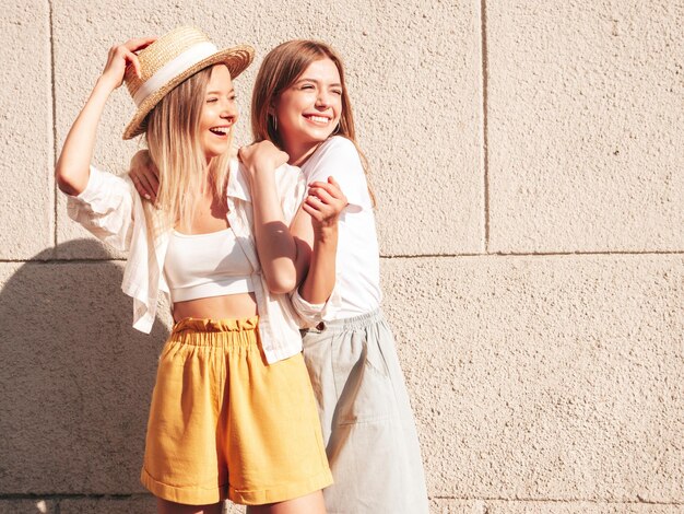 Dos jóvenes hermosas mujeres hipster sonrientes con ropa de verano de moda. Mujeres sexy despreocupadas posando en la calle cerca de una pared blanca con sombrero Modelos puros positivos divirtiéndose al atardecer