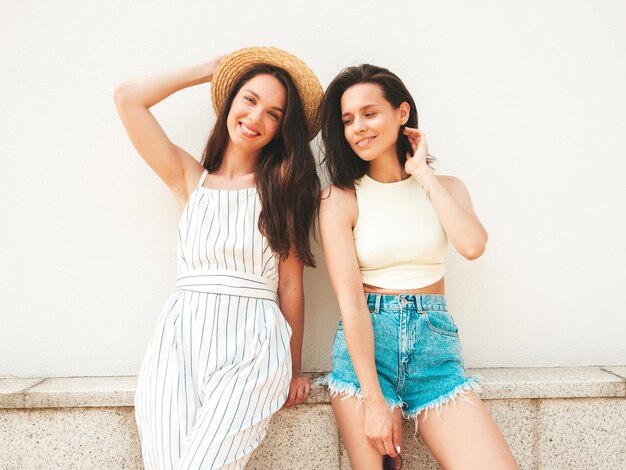 Dos jóvenes hermosas mujeres hipster sonrientes con ropa de verano de moda. Mujeres sexy despreocupadas posando en la calle cerca de la pared con sombrero.