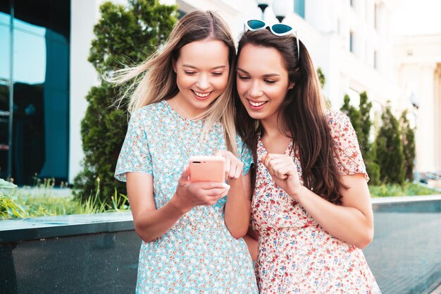 Dos jóvenes hermosas mujeres hipster sonrientes con ropa de verano de moda. Mujeres sexy y despreocupadas posando en la calle. Modelos positivos divirtiéndose abrazándose. Miran la pantalla del teléfono inteligente usando aplicaciones telefónicas.