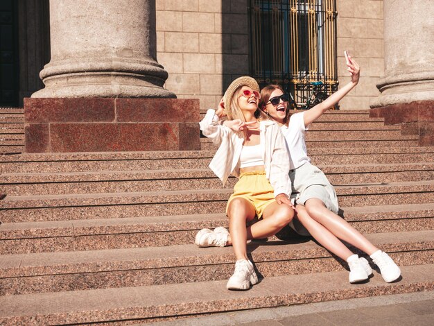 Dos jóvenes hermosas mujeres hipster sonrientes con ropa de verano de moda. Mujeres sexy y despreocupadas posando en la calle. Modelos puros positivos divirtiéndose al atardecer abrazándose y tomándose selfie. Sentadas en las escaleras.