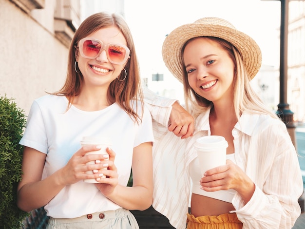 Dos jóvenes hermosas mujeres hipster sonrientes con ropa de verano de moda. Mujeres sexy y despreocupadas posando en la calle. Modelos puros positivos divirtiéndose al atardecer. Beben café y se comunican.