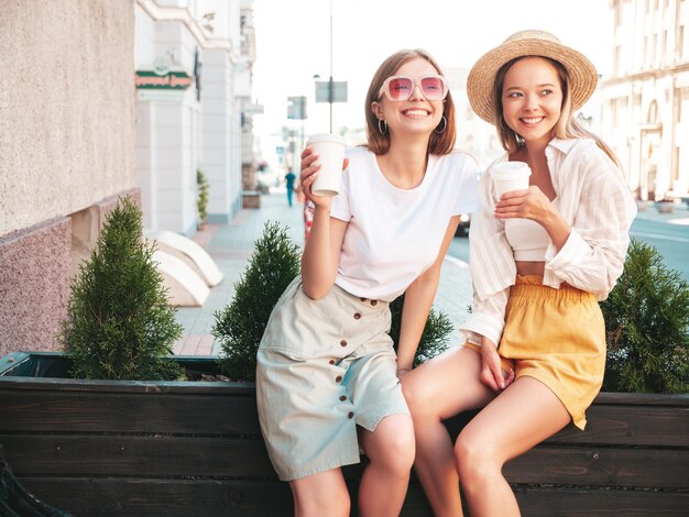 Dos jóvenes hermosas mujeres hipster sonrientes con ropa de verano de moda. Mujeres sexy y despreocupadas posando en la calle. Modelos puros positivos divirtiéndose al atardecer. Beben café o té en un vaso de plástico.