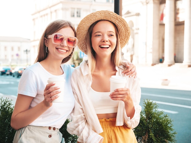 Dos jóvenes hermosas mujeres hipster sonrientes con ropa de verano de moda. Mujeres sexy y despreocupadas posando en la calle. Modelos puros positivos divirtiéndose al atardecer. Beben café o té en un vaso de plástico.