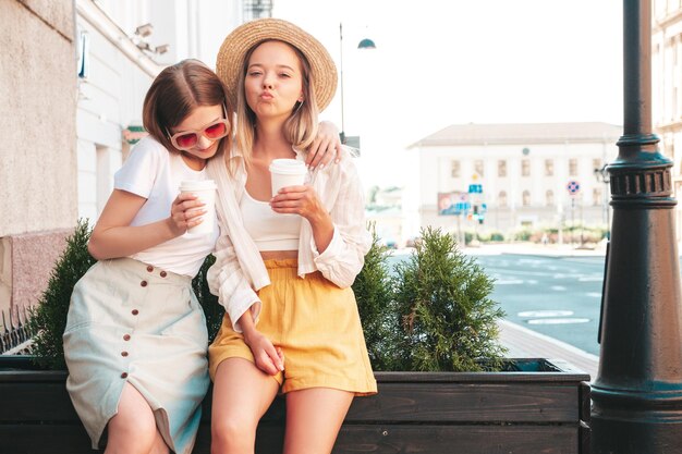 Dos jóvenes hermosas mujeres hipster sonrientes con ropa de verano de moda. Mujeres sexy y despreocupadas posando en la calle. Modelos puros positivos divirtiéndose al atardecer. Beben café o té en un vaso de plástico.