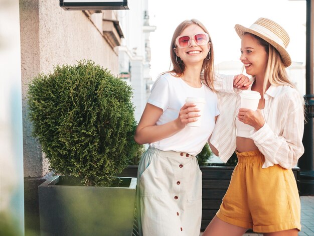 Dos jóvenes hermosas mujeres hipster sonrientes con ropa de verano de moda. Mujeres sexy y despreocupadas posando en la calle. Modelos puros positivos divirtiéndose al atardecer. Beben café o té en un vaso de plástico.