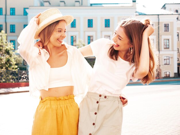 Dos jóvenes hermosas mujeres hipster sonrientes con ropa de verano de moda. Mujeres sexy despreocupadas posando en la calle con sombrero. Modelos puros positivos divirtiéndose al atardecer.