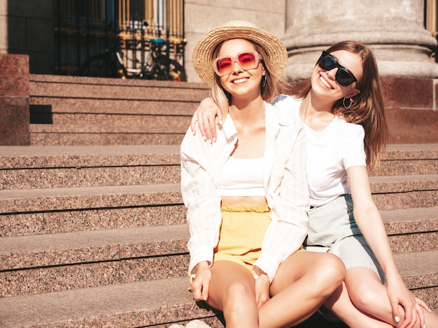 Dos jóvenes hermosas mujeres hipster sonrientes con ropa de verano de moda. Mujeres sexy despreocupadas posando en la calle con sombrero. Modelos puros positivos divirtiéndose al atardecer.