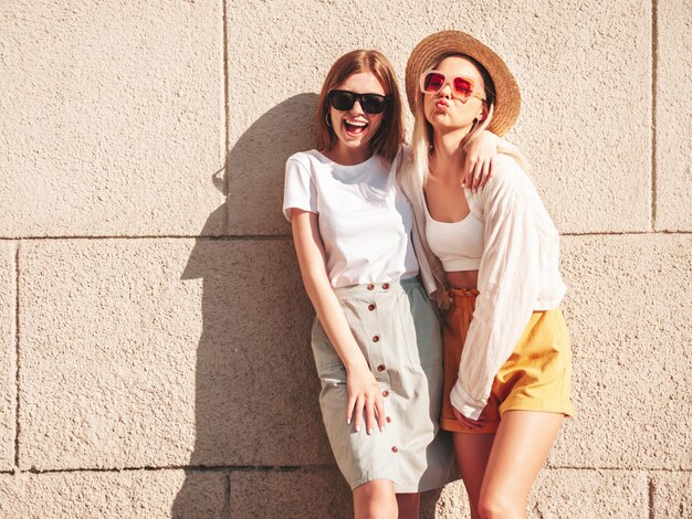 Dos jóvenes hermosas mujeres hipster sonrientes con ropa de verano de moda. Mujeres sexy despreocupadas posando cerca de una pared blanca en la calle. Modelos puros positivos divirtiéndose al atardecer abrazándose y volviéndose locos.