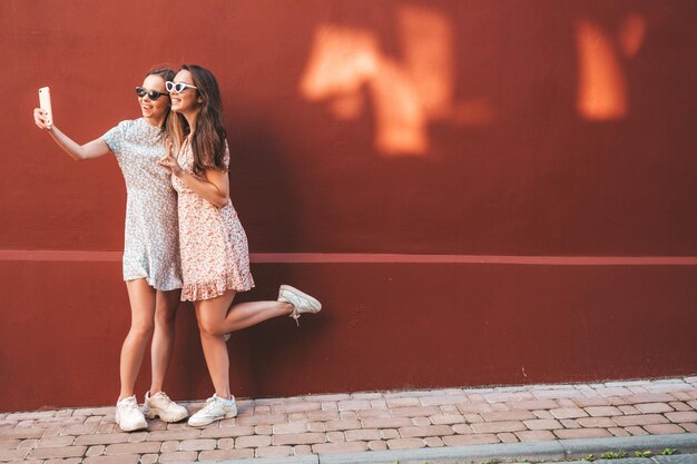 Dos jóvenes hermosas mujeres hipster sonrientes en vestidos de verano de moda Mujeres sexy despreocupadas posando en el fondo de la calle cerca de la pared roja Modelos puros positivos divirtiéndose al atardecer abrazándose Tomando selfie