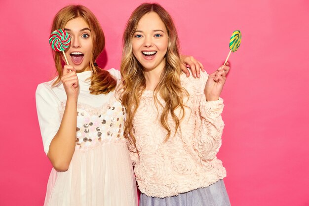 Dos jóvenes hermosas mujeres rubias hipster sonrientes en ropa de moda de verano. Mujeres calientes despreocupadas que presentan cerca de la pared rosada. Modelos divertidos positivos abrazándose con piruleta