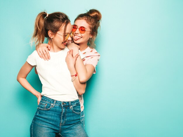 Dos jóvenes hermosas rubias sonrientes chicas hipster en ropa de jeans de moda hipster de verano. Mujeres despreocupadas sexy posando junto a la pared azul. Modelos modernos y positivos divirtiéndose en gafas de sol