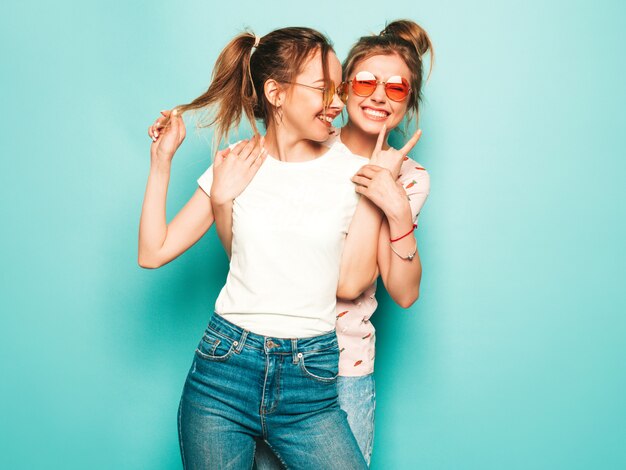 Dos jóvenes hermosas rubias sonrientes chicas hipster en ropa de jeans de moda hipster de verano. Mujeres despreocupadas sexy posando junto a la pared azul. Modelos modernos y positivos divirtiéndose en gafas de sol