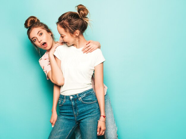 Dos jóvenes hermosas rubias sonrientes chicas hipster en ropa de jeans de moda hipster de verano. Mujeres despreocupadas sexy posando junto a la pared azul. Modelos modernos y positivos divirtiéndose