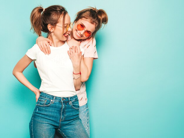 Dos jóvenes hermosas rubias sonrientes chicas hipster en ropa de jeans de moda hipster de verano. Mujeres despreocupadas sexy posando junto a la pared azul. Modelos modernos y positivos divirtiéndose