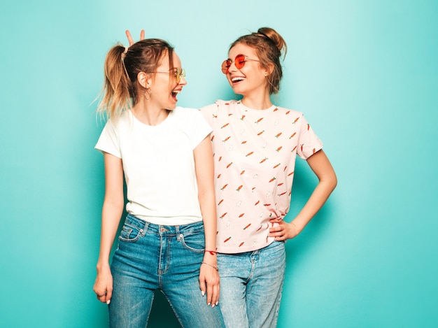 Dos jóvenes hermosas rubias sonrientes chicas hipster en ropa de jeans de moda hipster de verano. Mujeres despreocupadas sexy posando junto a la pared azul. Modelos modernos y positivos divirtiéndose
