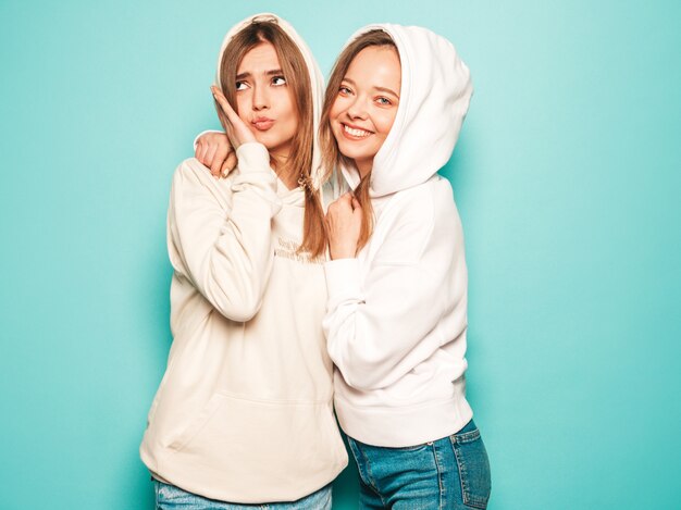 Dos jóvenes hermosas rubias sonrientes chicas hipster en ropa de moda con capucha de verano. Mujeres despreocupadas sexy posando junto a la pared azul. Modelos modernos y positivos divirtiéndose