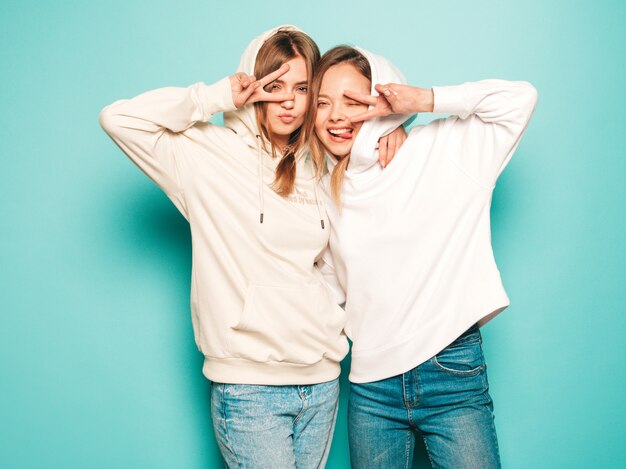 Dos jóvenes hermosas rubias sonrientes chicas hipster en ropa de moda con capucha de verano. Mujeres despreocupadas sexy posando junto a la pared azul. Modelos modernos y positivos muestran signos de paz en gafas de sol