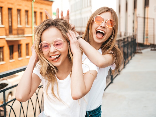 Dos jóvenes hermosas rubias sonrientes chicas hipster en ropa de moda verano blanco. Modelos positivos divirtiéndose en gafas de sol. Usando los dedos como orejas de conejo.