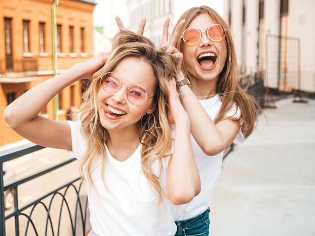 Dos jóvenes hermosas rubias sonrientes chicas hipster en ropa de moda verano blanco. Modelos positivos divirtiéndose en gafas de sol. Usando los dedos como orejas de conejo.